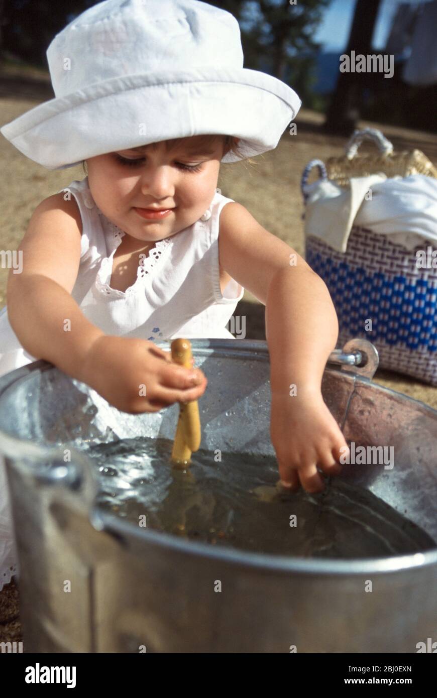 Kleines Mädchen spielt mit einem Eimer mit klarem Wasser und einigen Wäscheklammern, während Mutter die Wäsche im sonnigen Hof hängt - Stockfoto