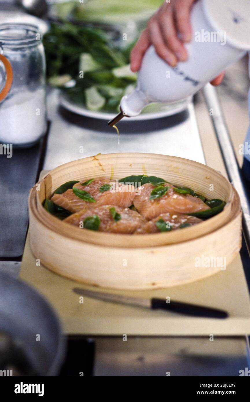 In einem chinesischen Bambusdampfer wird Olivenöl über Lachsfilets mit Basilikumblättern tröpfelt. - Stockfoto