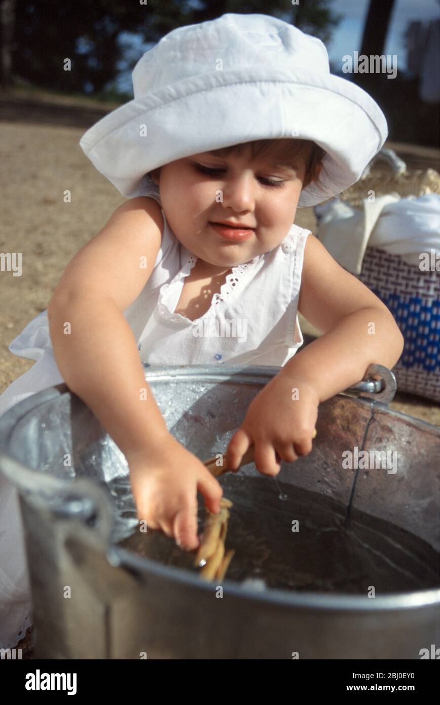 Kleines Mädchen, das mit einem Eimer mit klarem Wasser und einigen Wäscheklammern blasset, während Mutter die Wäsche im sonnigen Hof hängt - Stockfoto