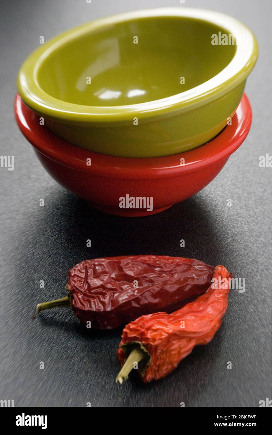 Zwei helle Melaminschalen auf dunkler Oberfläche mit zwei roten scharfen Chilischoten gestapelt - Stockfoto