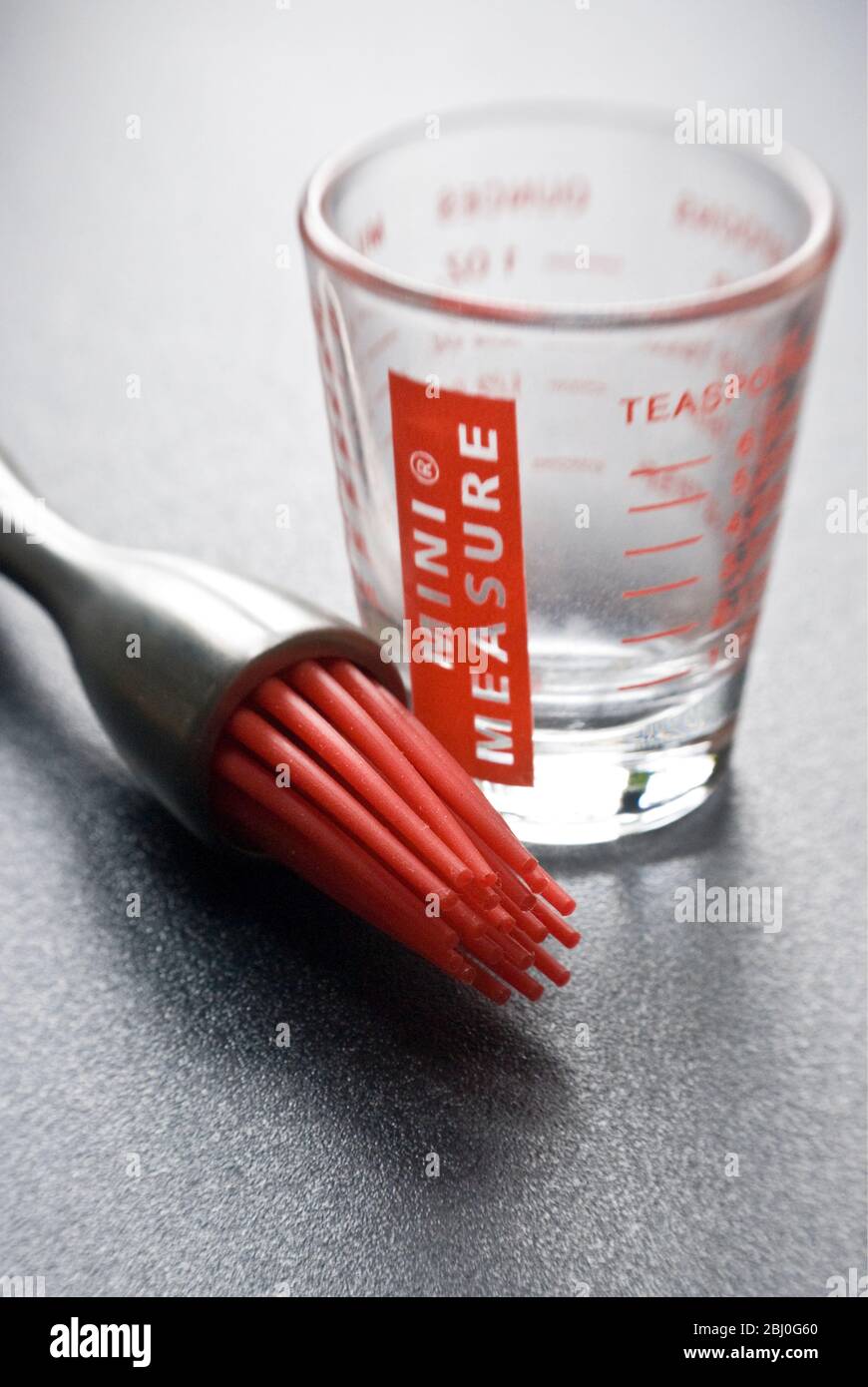 Moderne Teigbürste mit roten Silikon-Borsten mit einem kleinen Messglas auf dunkler strukturierter Oberfläche - Stockfoto