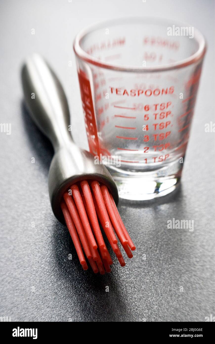Moderne Teigbürste mit roten Silikon-Borsten mit einem kleinen Messglas auf dunkler strukturierter Oberfläche - Stockfoto