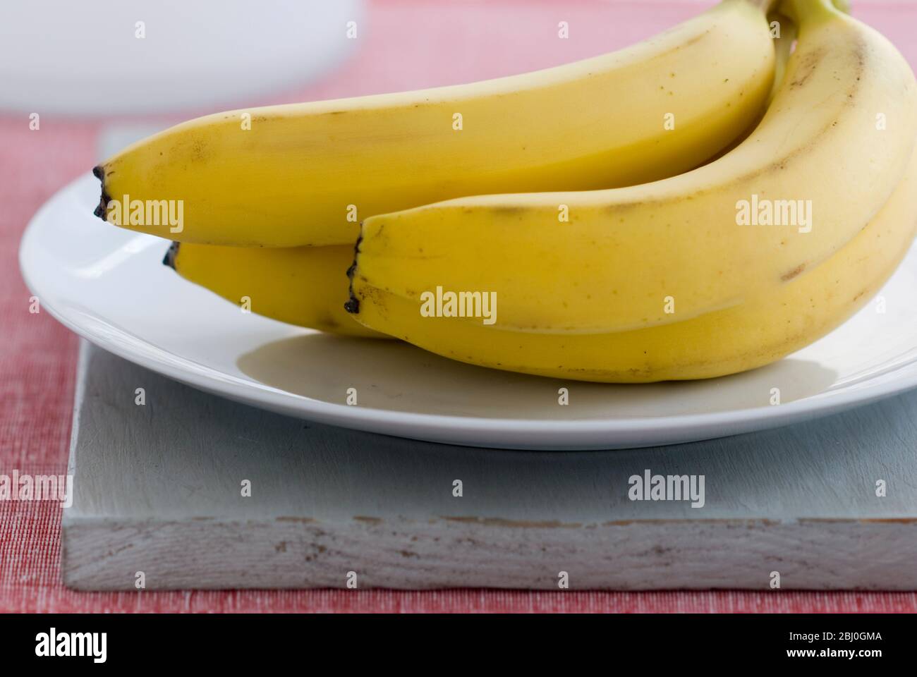 Bananenstrauß auf weißem Teller, auf grauem Brett, mit rotem Hintergrund - Stockfoto