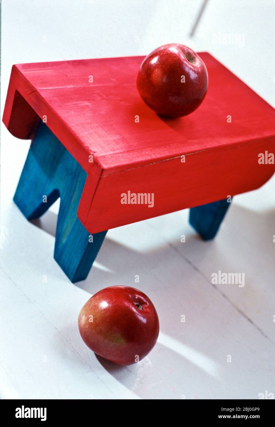 Zwei leuchtend rote Äpfel mit hell bemalten Fußhocker auf weißem Holzboden. - Stockfoto