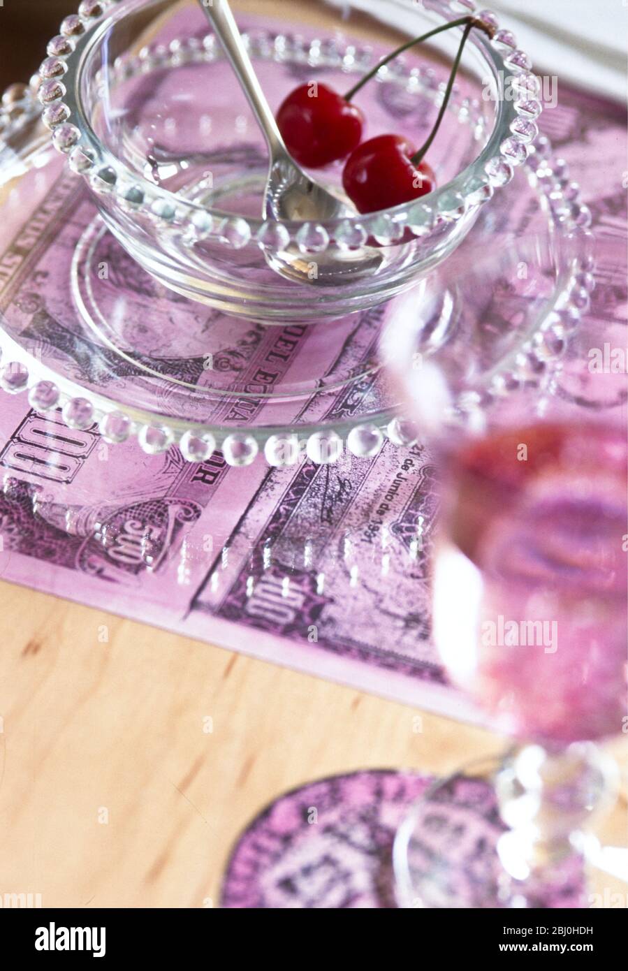 Ein Paar Kirschen in einer Glasschale mit Platzmatte aus vergrößerten Fotokopien alter Banknoten - Stockfoto