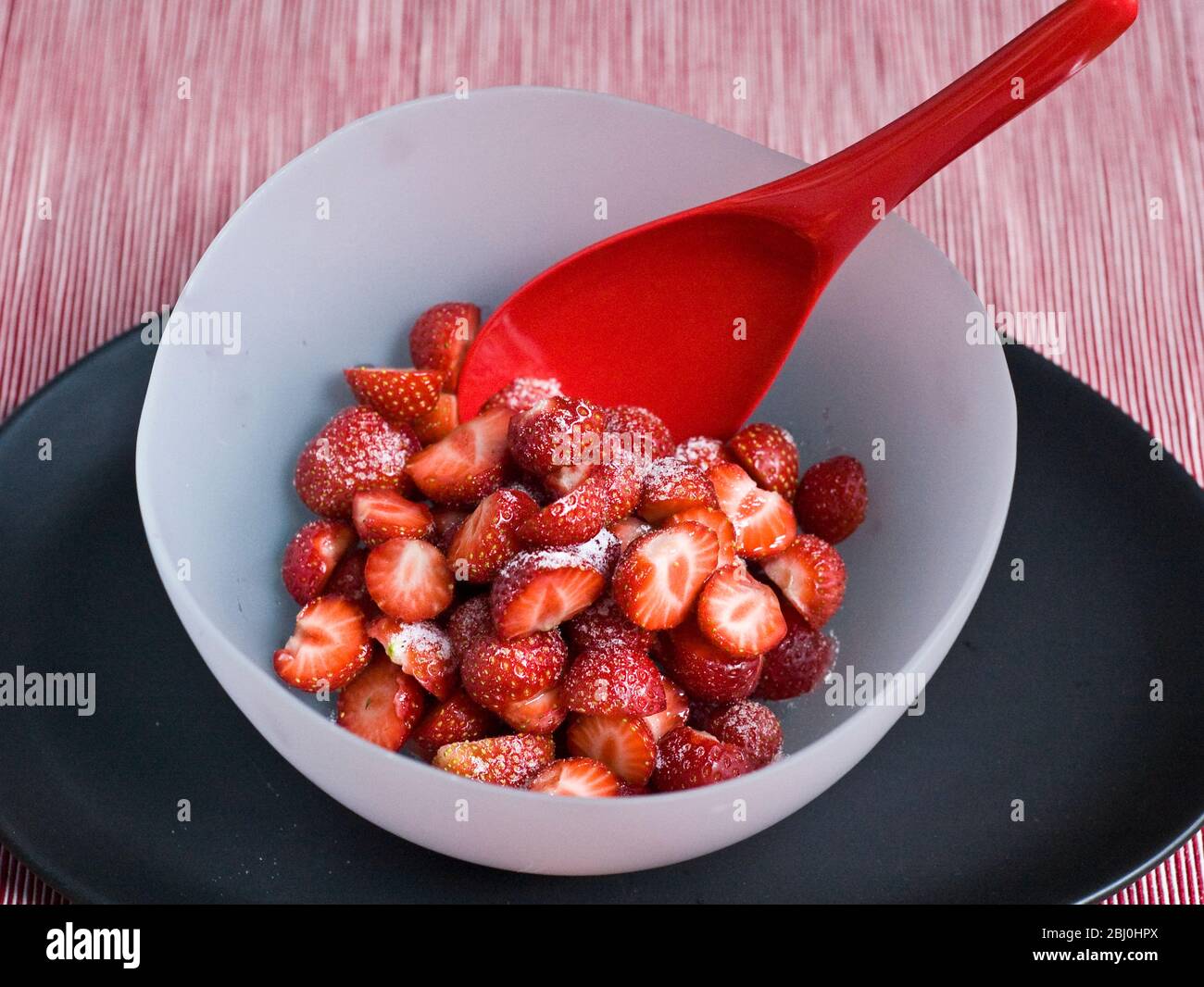 Erdbeeren, geschält und halbiert, mit Gießzucker bestäubt, in einer Schüssel aus mattem Glas auf schwarzem Teller mit hellrotem Melaminlöffel serviert. - Stockfoto