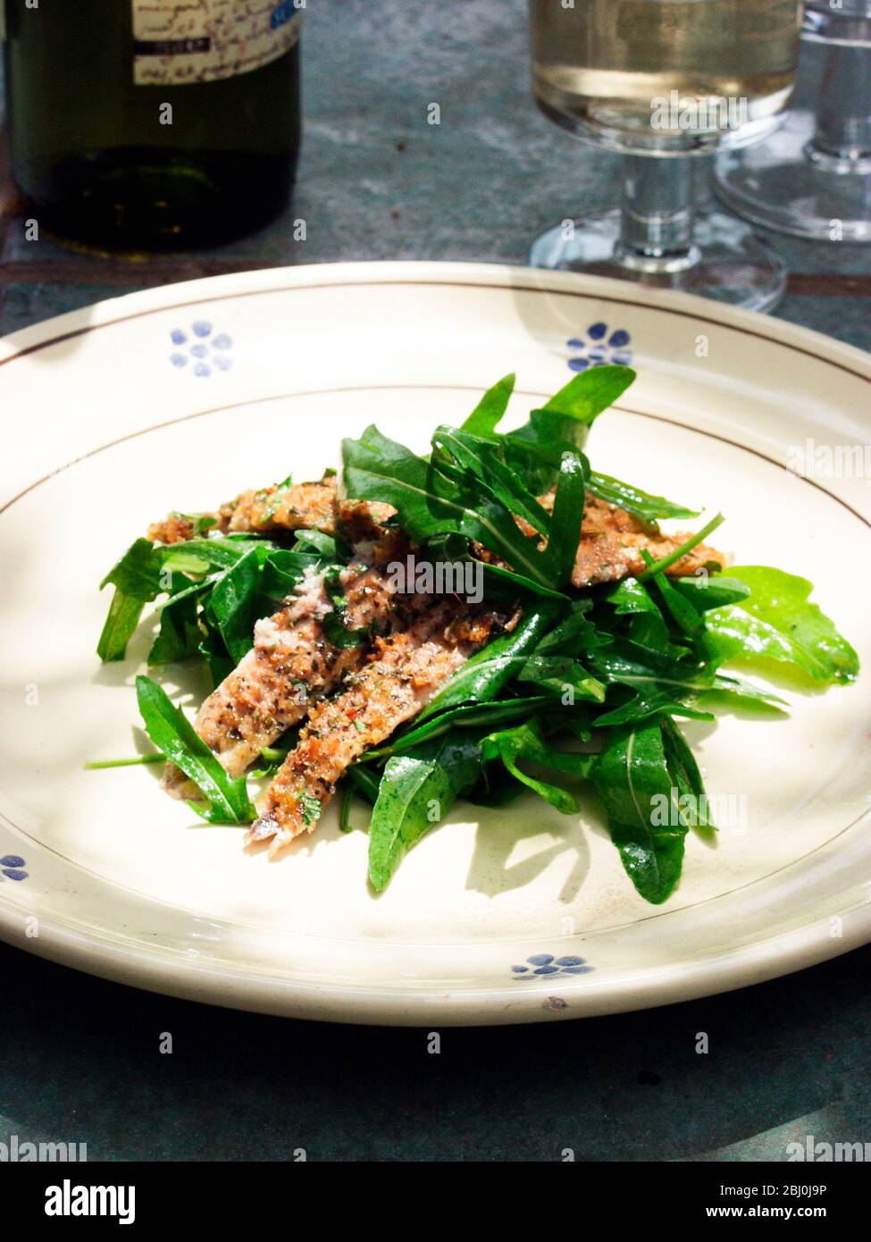 Vorspeise aus pangebratenen Sardellenfilets mit Rucola-Salat, serviert im Freien, bei Sonnenschein mit Wein - Stockfoto