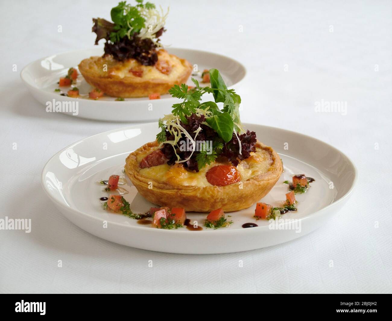 Kleine herzhafte Party Quiches mit dekorativer Beilage von Baby-Blatt-Salat und umgeben von fein gehackten Tomaten und Kräutern mit Öl und Balsamico-Rebe Stockfoto