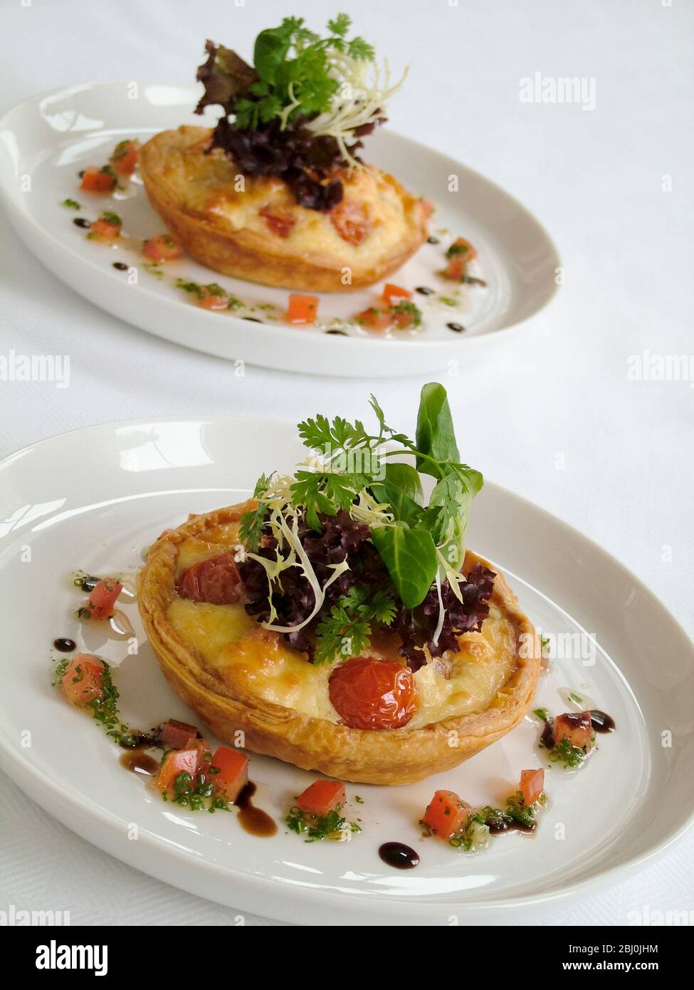 Kleine herzhafte Party Quiches mit dekorativer Beilage von Baby-Blatt-Salat und umgeben von fein gehackten Tomaten und Kräutern mit Öl und Balsamico-Rebe Stockfoto