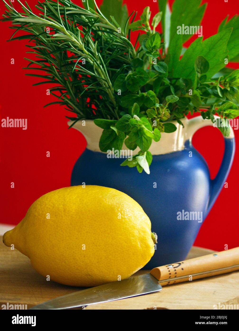 Zitrone auf Schneidebrett mit Messer und Bund gemischter Gartenkräuter in blauem Krug vor rotem Hintergrund - Stockfoto