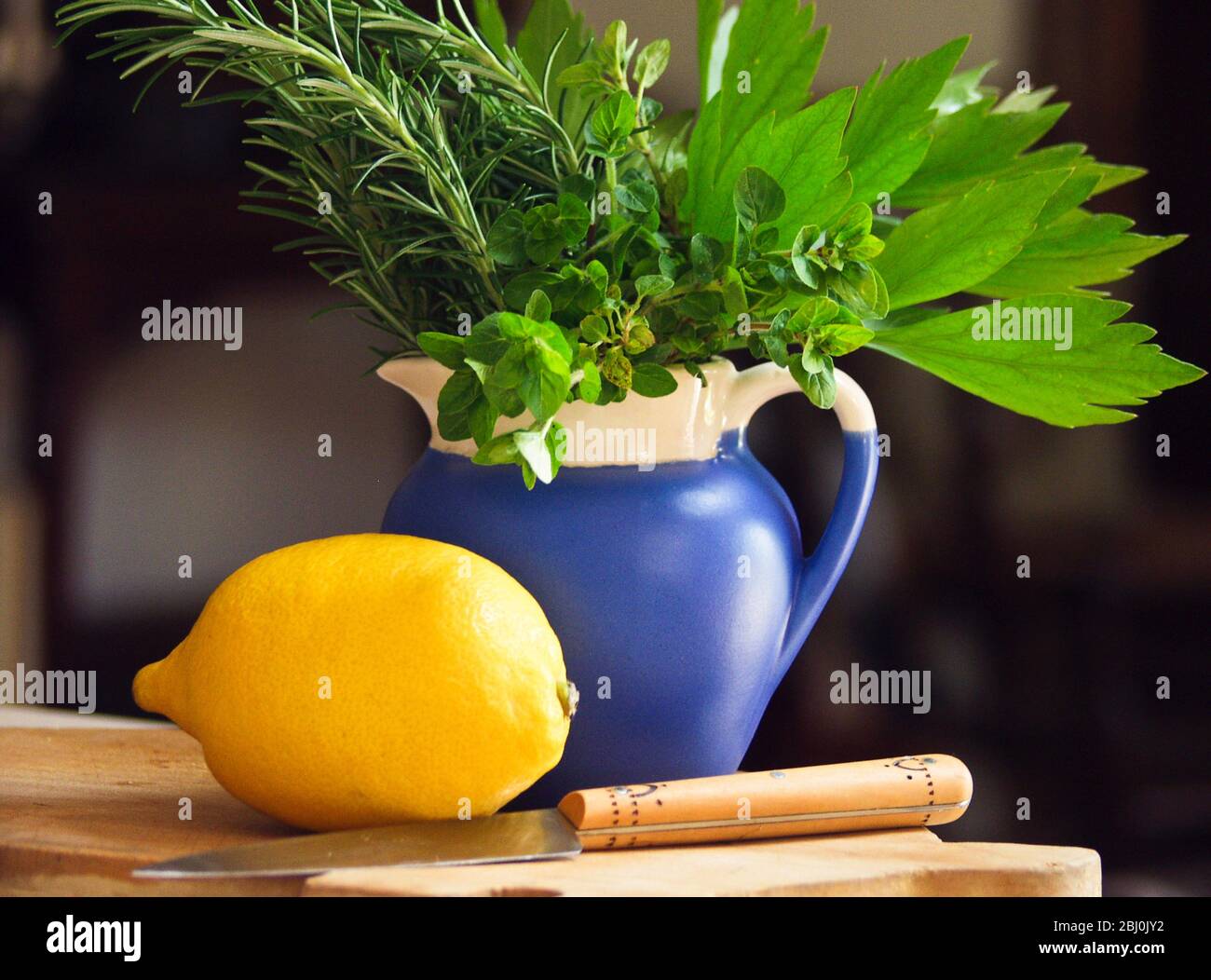 Zitrone auf Schneidebrett mit Messer und Bund gemischter Gartenkräuter in blauem Krug vor dunklem Hintergrund - Stockfoto