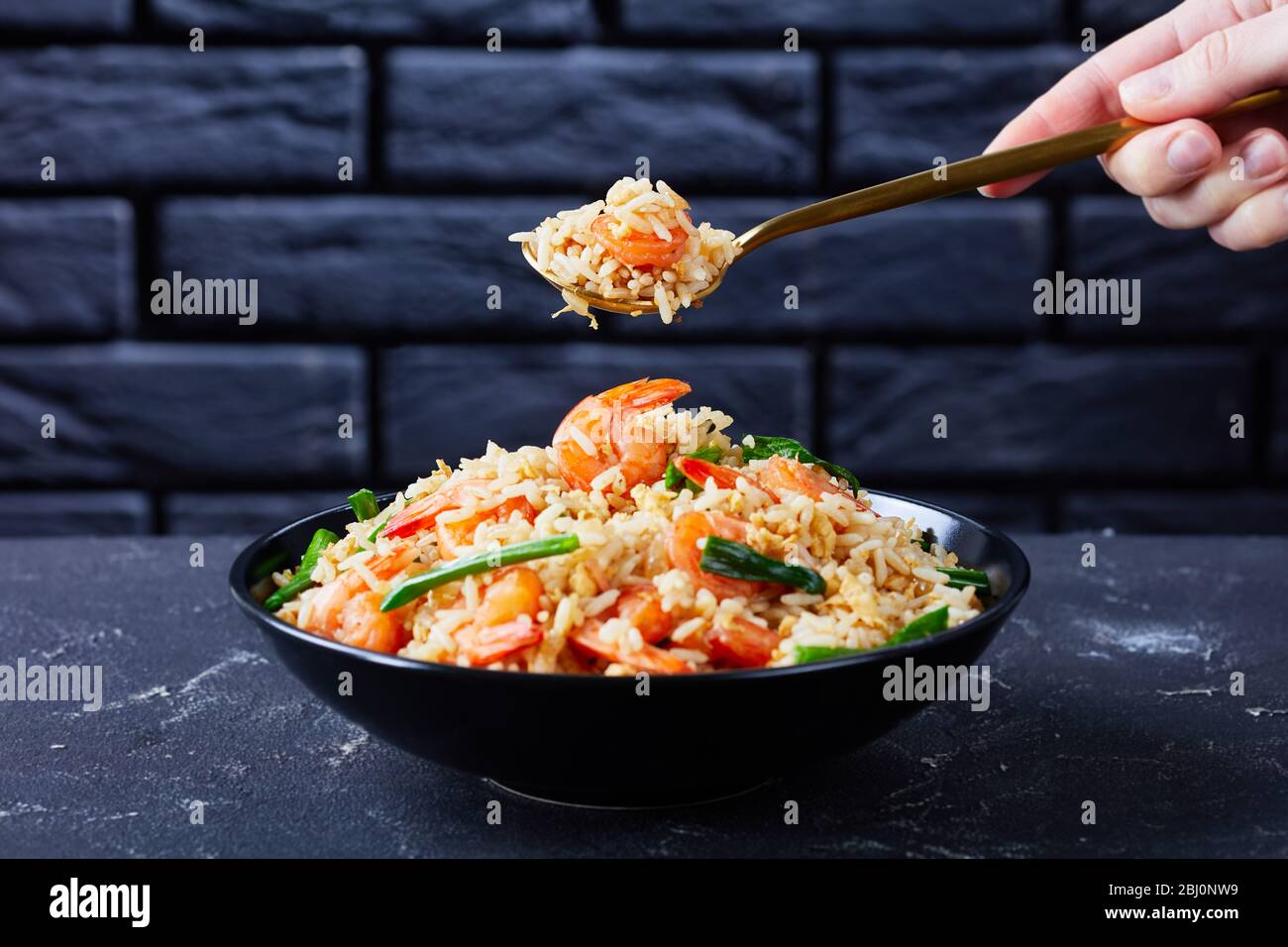 Frauenhand hält den Löffel mit asiatischem gebratenem Jasminreis mit Garnelen Rührei und Frühlingszwiebeln mit thai-Sauce auf dunklem Betongrund Stockfoto