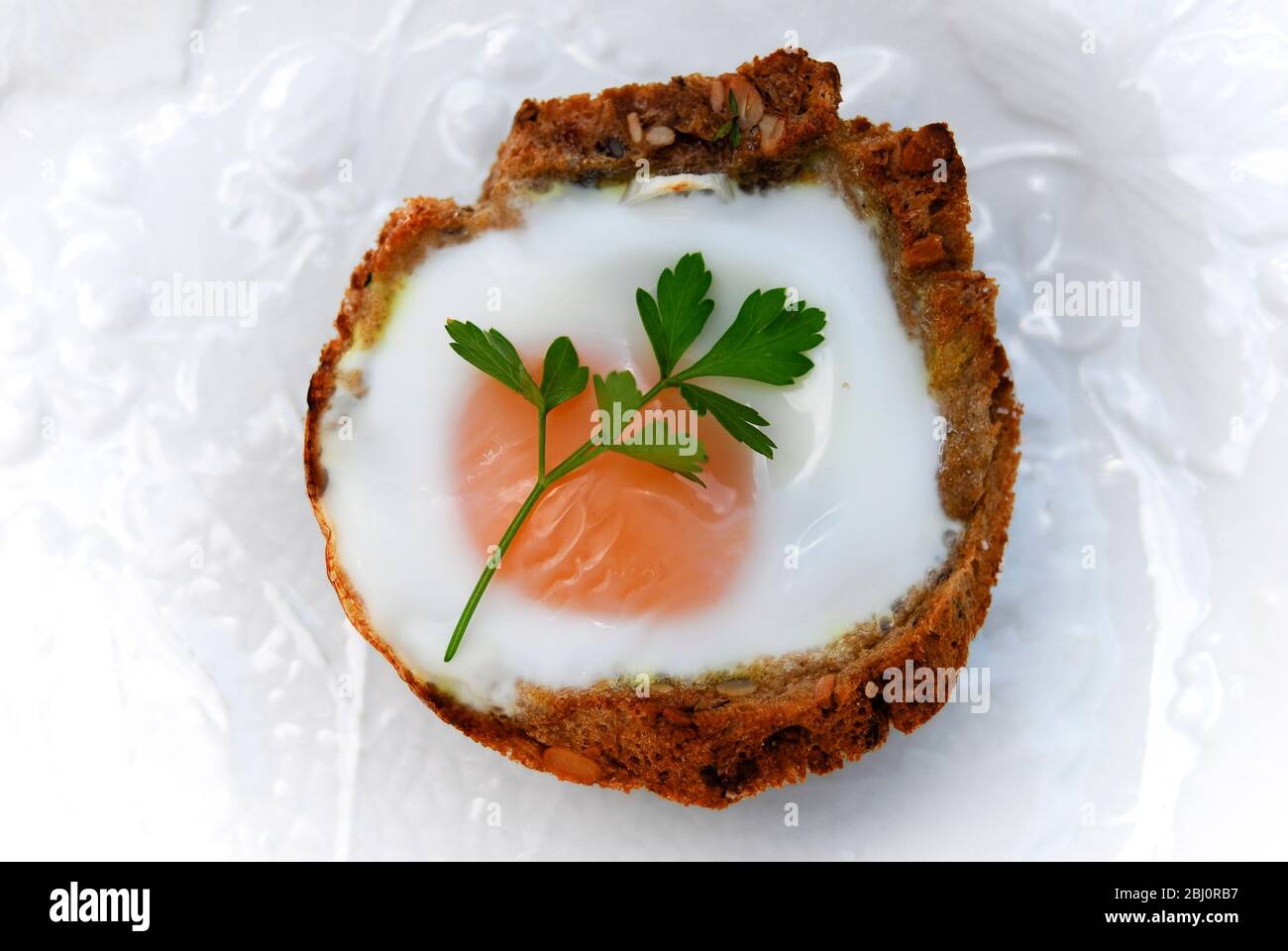 Ein Ei, das in einem Fall von gebuttertem Brot gebacken wurde, das auf Muffin-Form geschnitten wurde, garniert mit flacher Petersilie - Stockfoto