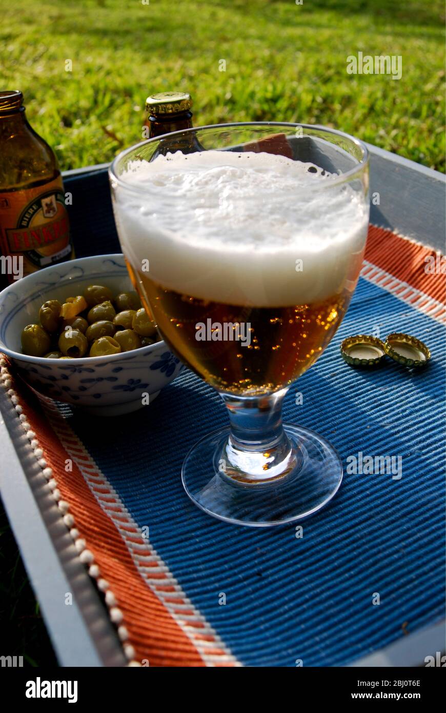 Ein Glas Bier mit einem guten Schaumkessel und einer Schale Oliven, auf einem Tablett auf dem Gras - Stockfoto