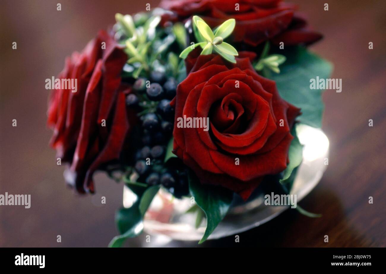 Opulente Tischdekoration aus samtigen roten Rosen und Laub in Glasvase auf dunklem Holztisch - Stockfoto