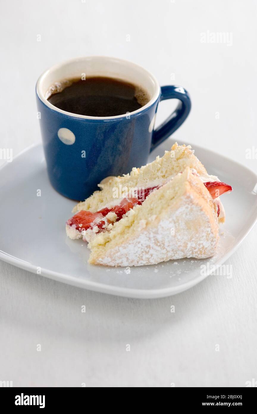 Stück Biskuitkuchen mit Puderzucker bestäubt und mit Erdbeeren und Sahne auf Teller mit kleinen blau-weiß gefleckten Becher schwarzen Kaffees gefüllt - Stockfoto