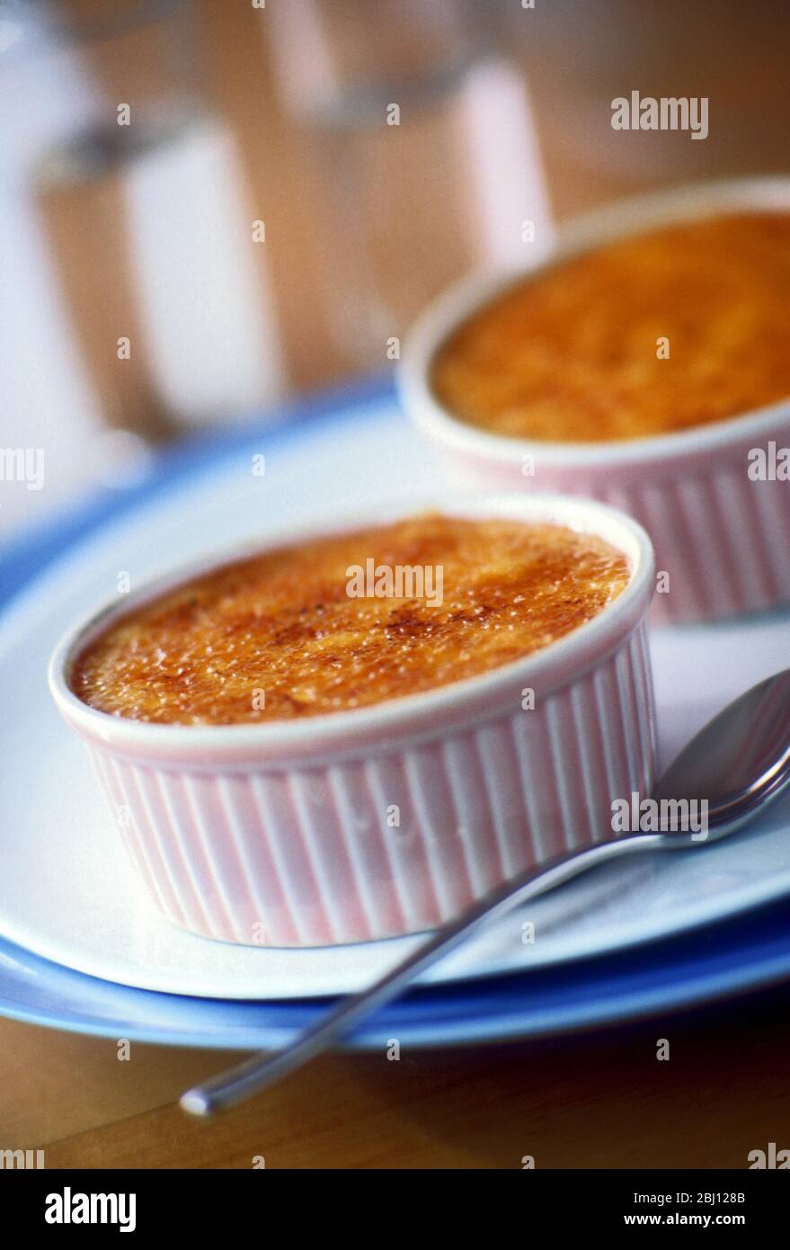 Creme brulee in zwei rosa Ramekins auf weißem Teller mit Gläsern dahinter - Stockfoto