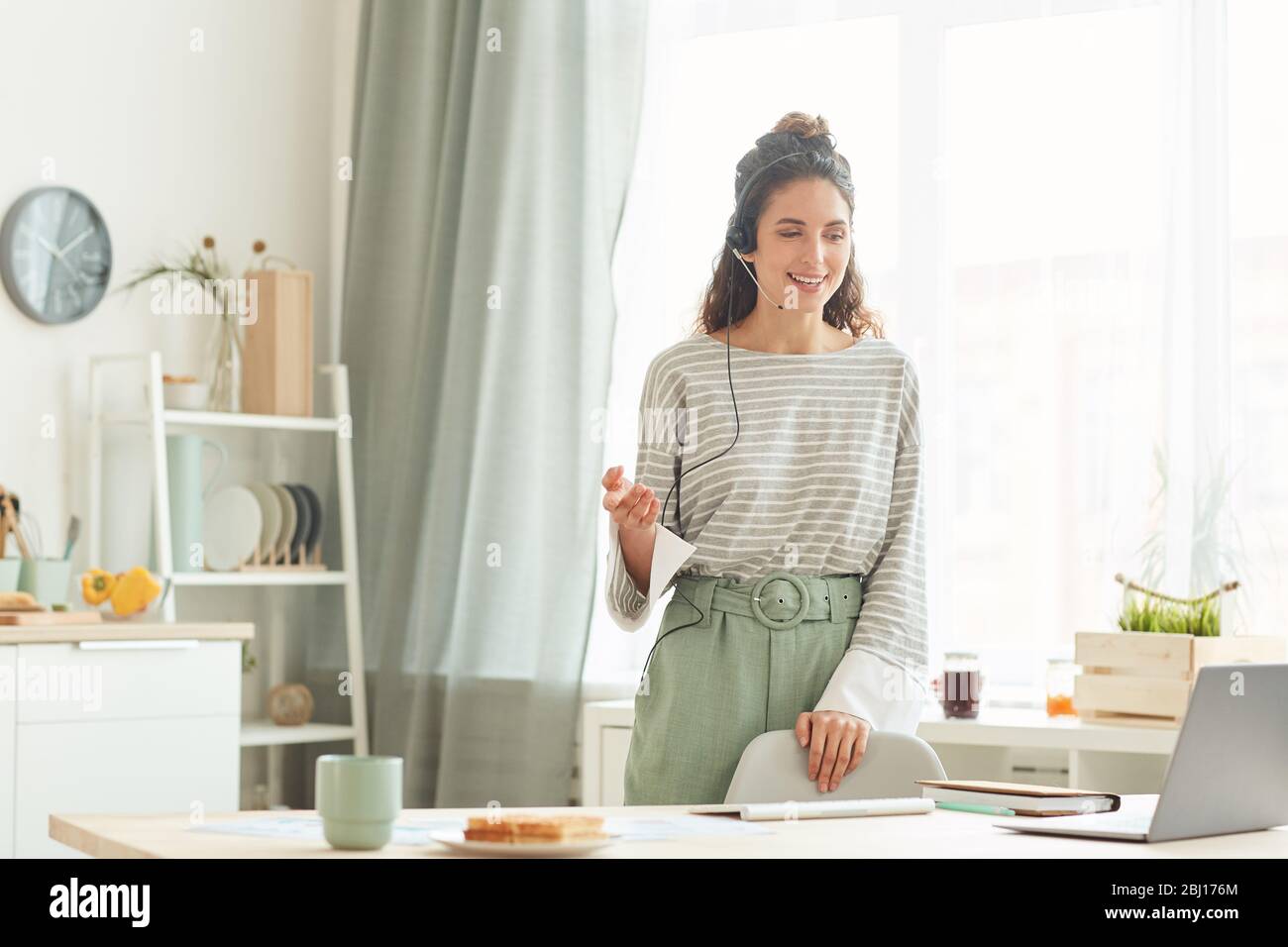 Junge kaukasische Frau mit lässigem Outfit stehend in der Küche zu Hause im Gespräch mit ihren Kollegen während der Online-Sitzung Stockfoto