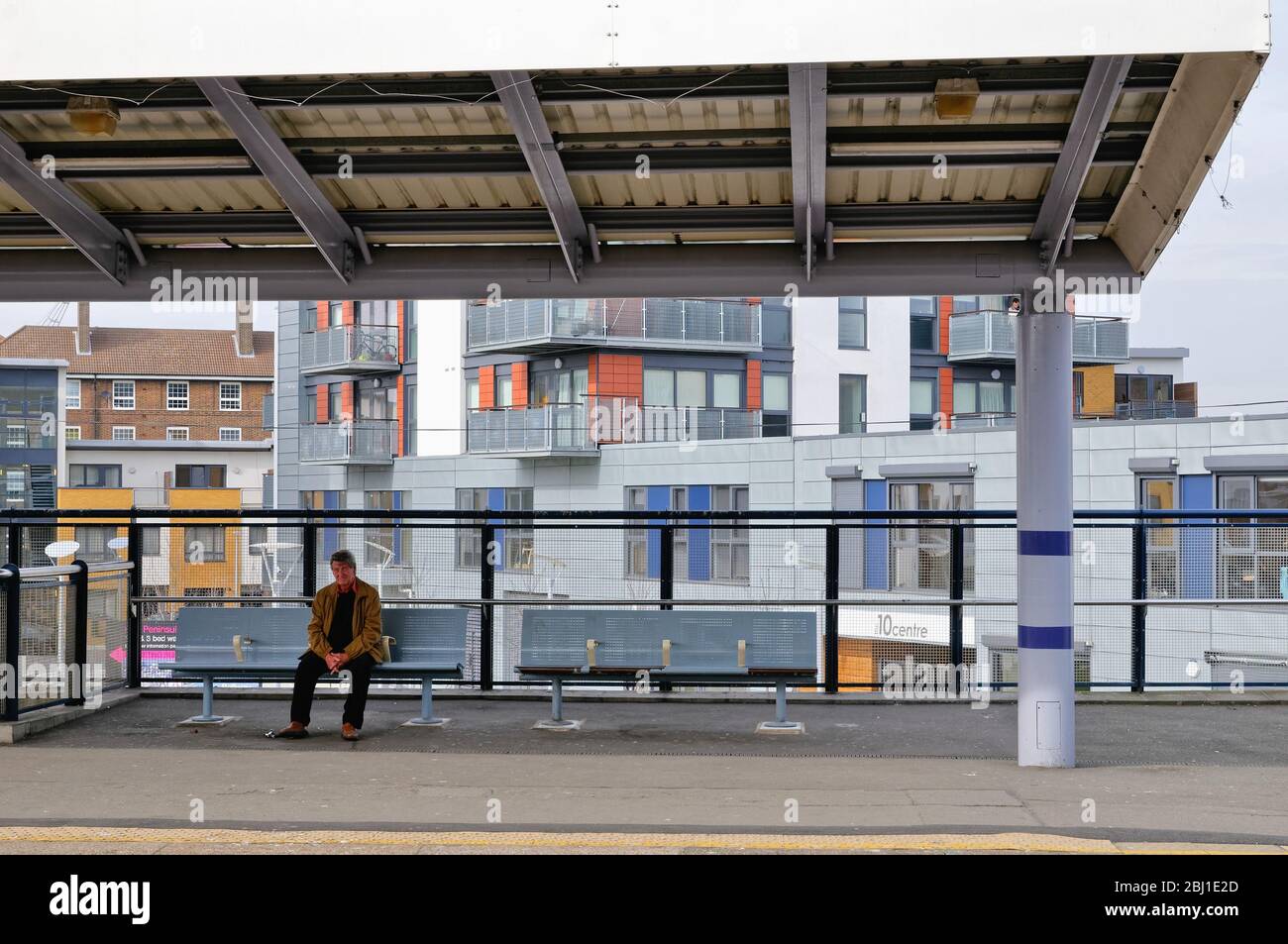 Ein Mann mittleren Alters, der auf dem Bahnsteig der Greenwich Hauptbahnstation sitzt und auf einen Zug mit modernen Apartments hinter London England UK wartet Stockfoto