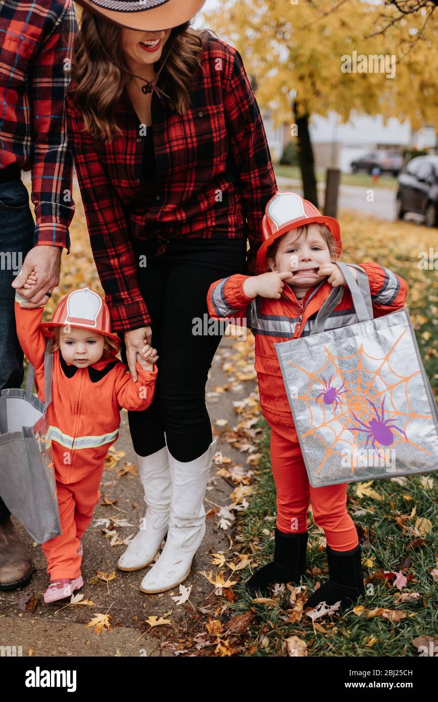 Eine Familie mit zwei Kindern, die als Feuerwehrleute für Halloween gekleidet sind, spazieren durch einen blattbesäten Park. Stockfoto