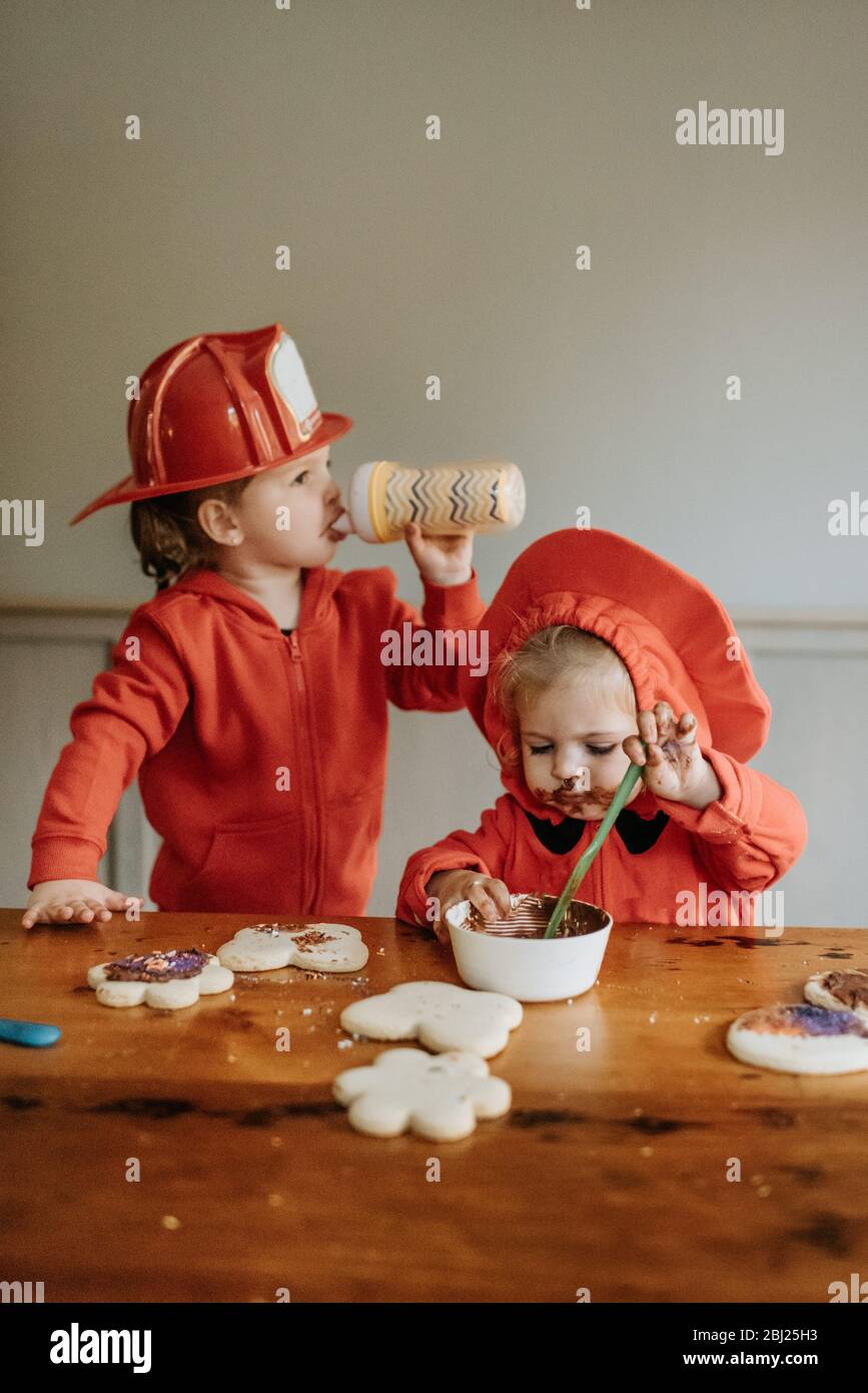 Zwei Kinder, die als Feuerwehrleute an einem Tisch gekleidet sind, dekorieren Kekse mit Pralinen und Streuseln. Stockfoto