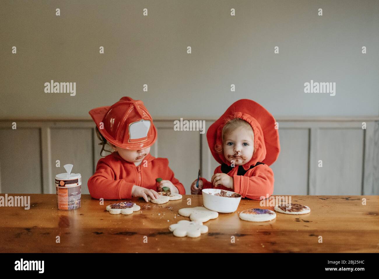 Zwei Kinder, die als Feuerwehrleute an einem Tisch gekleidet sind, dekorieren Kekse mit Pralinen und Streuseln. Stockfoto