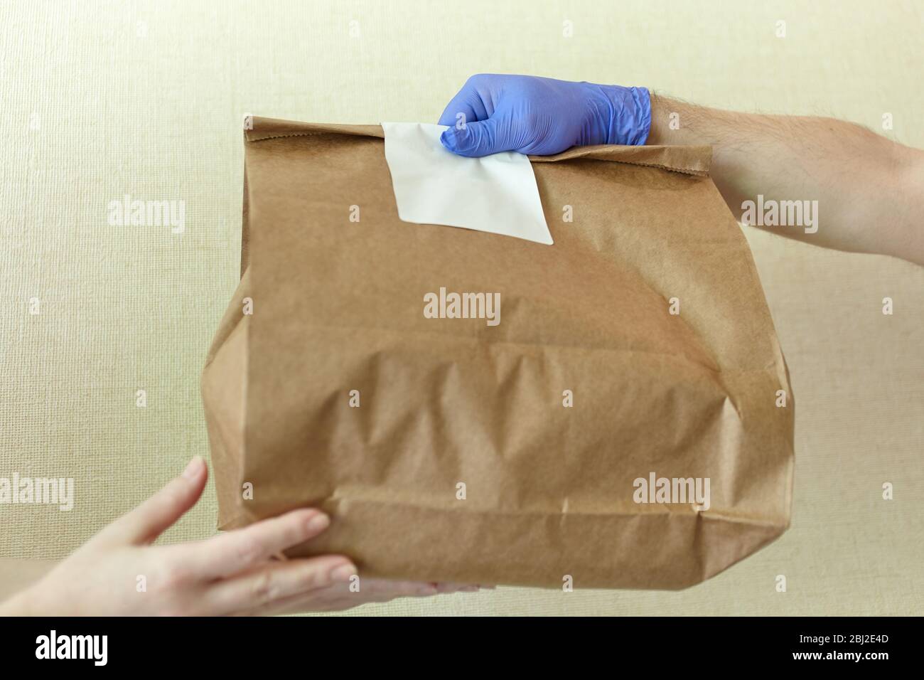 Männliche Hände in blauem Latex Handschuh gibt Papiertüte an weibliche Hände. Nahaufnahme der männlichen Hände in Handschuhen halten braunen Bastelpapierbeutel mit leeren weißen Aufkleber. D Stockfoto