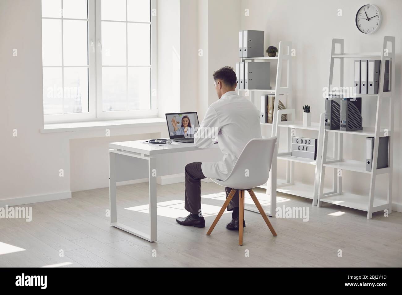 Arzt online. Der männliche Arzt arbeitet, während er Videoanruf mit dem Patienten mit einem Bast-Schuh spricht, während er am Schreibtisch im Büro der Klinik sitzt. Stockfoto