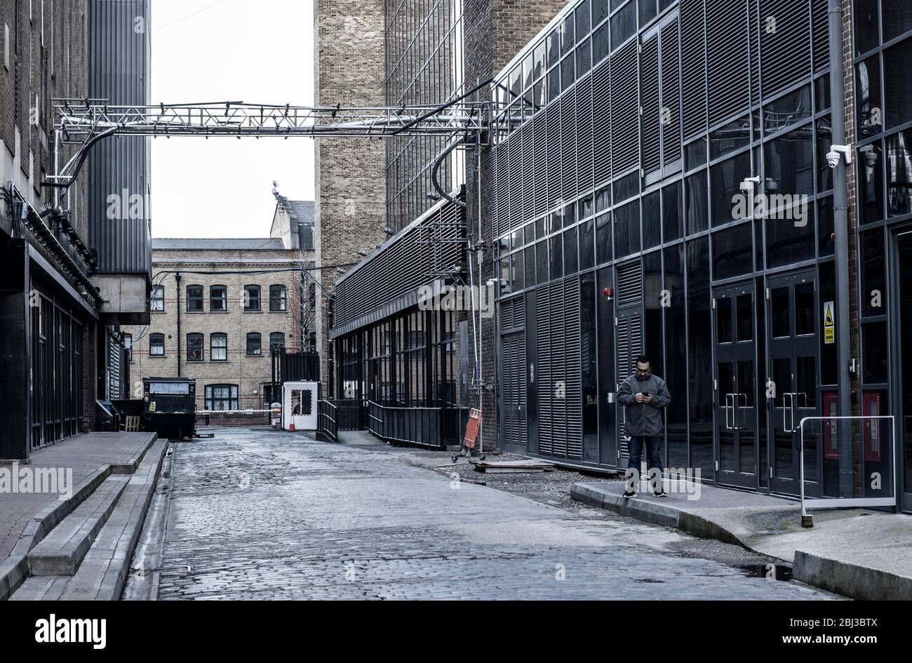 Ein Mann in grauer Kleidung steht einsam inmitten eines Industrieanwesens in East London, wo die Gebäude die gleichen Farben wie seine Kleidung haben. Stockfoto