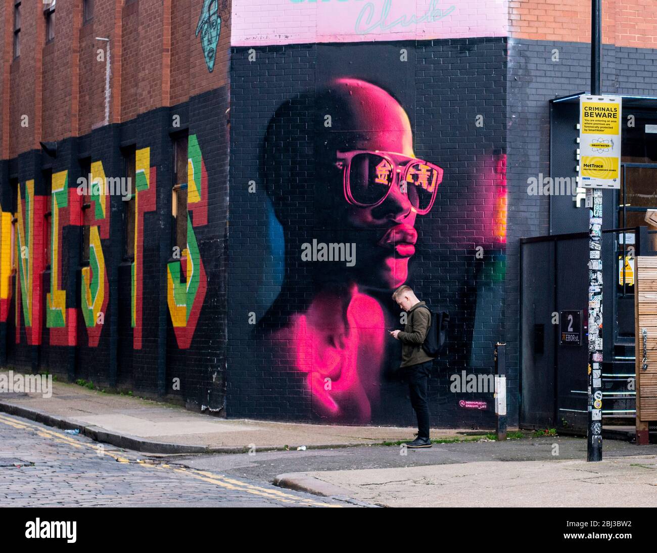 Ein junger Mann steht vor einem riesigen Graffiti-Kunstwerk, dessen Kopf das Kinn eines großen gemalten Mannes in Shoreditch in East London berührt. Stockfoto