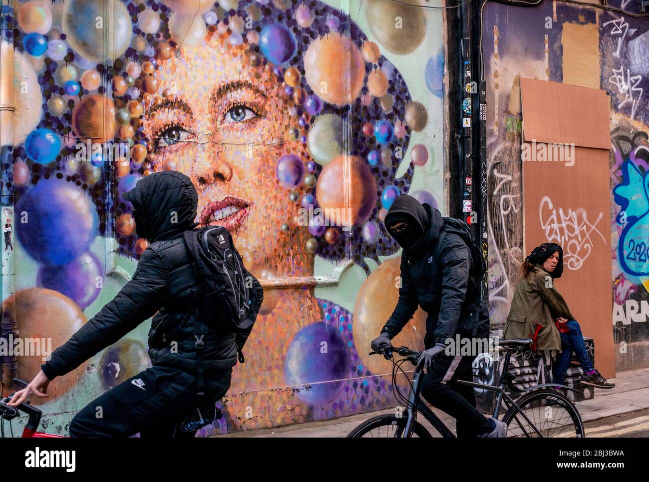 Zwei junge Hooded Cycle vorbei an einem riesigen Stück Graffiti-Kunst, während eine Dame in der Nähe sitzt und die Kamera betrachtet. Stockfoto