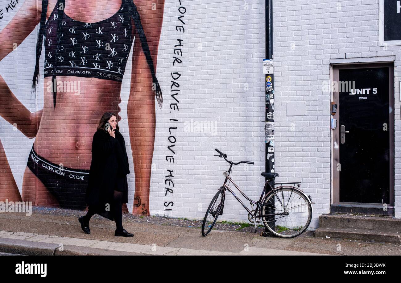 Eine in schwarzen Spaziergängen gekleidete Dame vorbei an einem Graffiti-Calvin Klein-Werbespot neben einer schwarzen Tür und einem schwarzen Fahrrad in Shoreditch im Osten Londons. Stockfoto