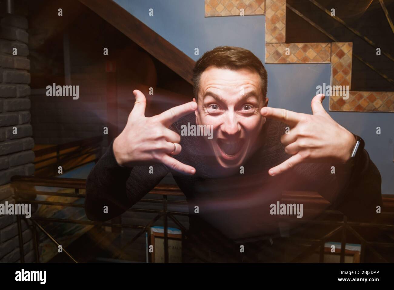 Das Gesicht eines Mannes mit Emotionen. Junger emotionaler Mann. Menschliche Emotionen, Gesichtsausdruck Konzept. Menschenhände machen ein Zeichen von Hörnern. Stockfoto