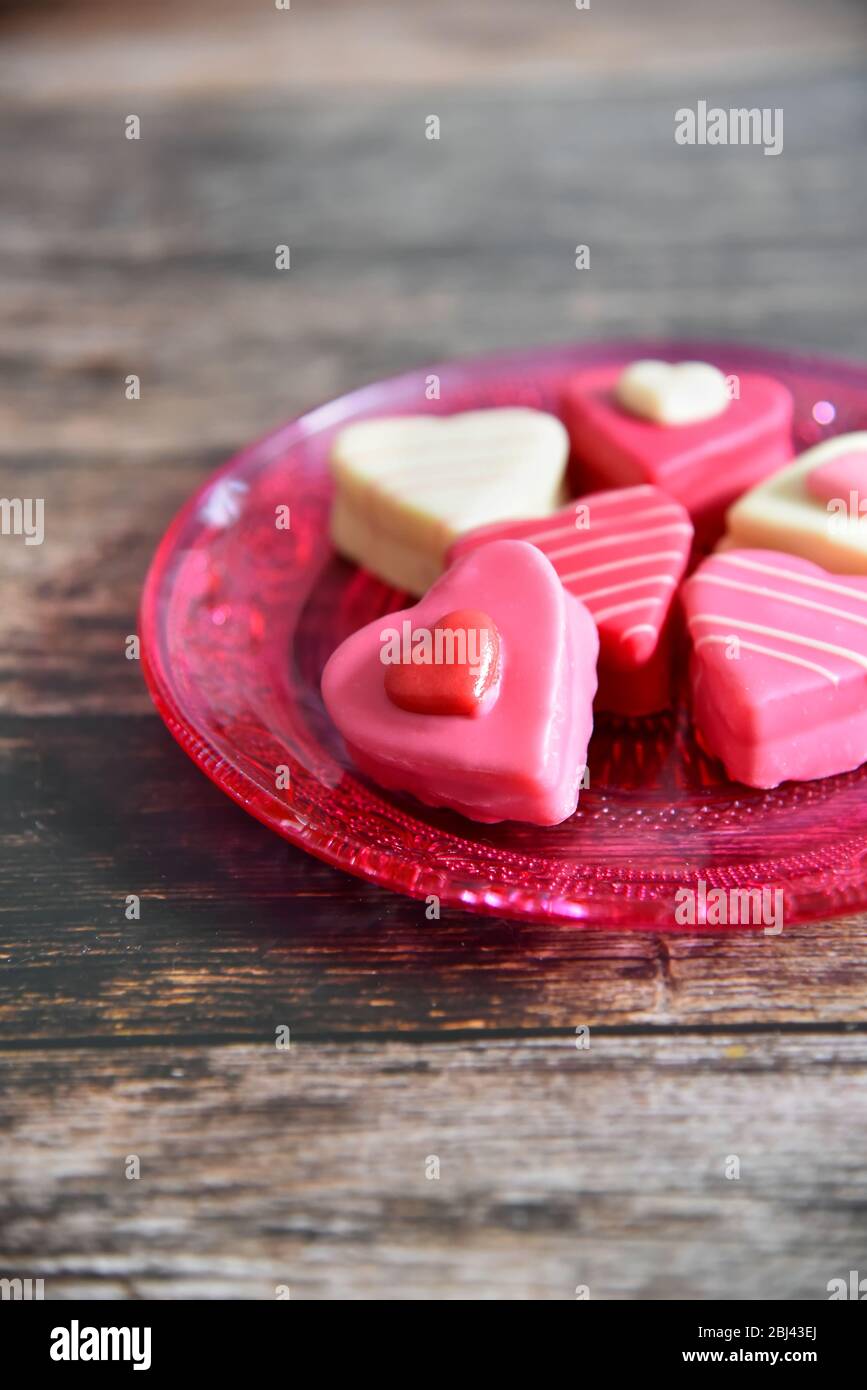 Herzförmige, farbenfrohe Pralinen auf einem rosa Glasteller mit Ornamentmuster. Dunkler Holztisch Hintergrund. Liebe und Grußkarte Konzept. Stockfoto