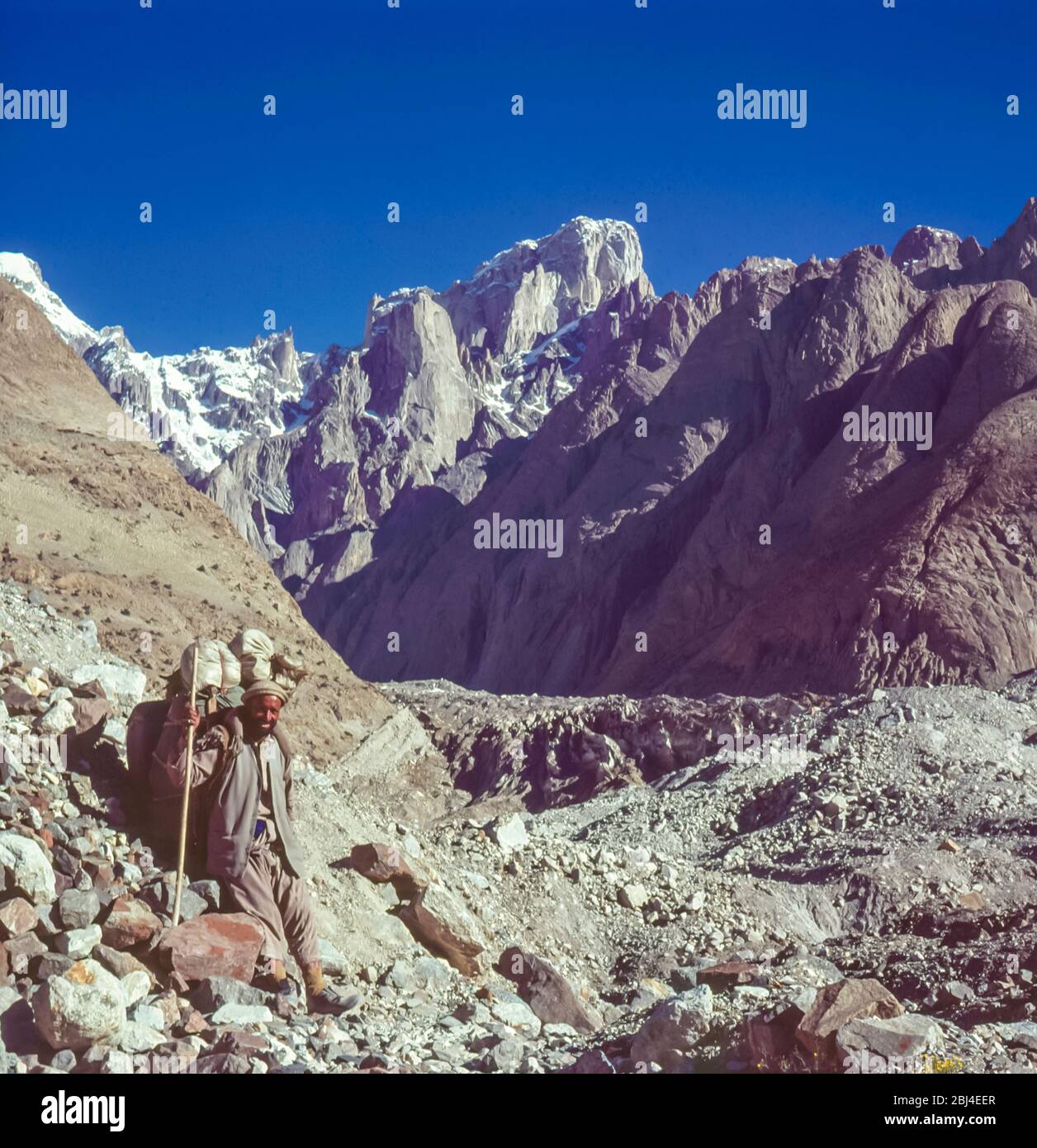 Pakistan, nördliche Gebiete der Karakorum-Berge. Ein einsames Balti-Portier macht eine Pause auf den Sitten des Baltoro-Gletschers mit dem Gipfel von Paiju in der Ferne. Stockfoto