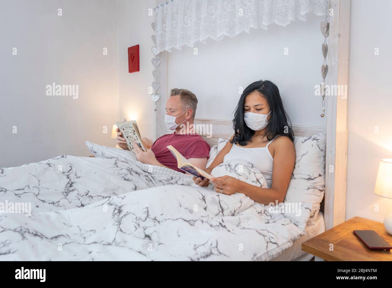Ein Paar im Bett trägt Gesichtsmasken, die Bücher lesen, während es sich selbst isoliert, während es sich im Lockdown versperrt Stockfoto