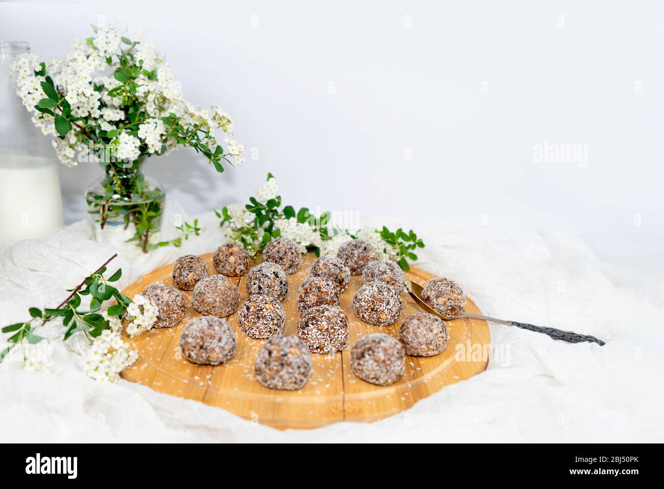 Protein Erdnussbutter Energie Bisse oder Energiebälle. Hausgemachte rohe Schokoladentrüffel mit Nüssen und in Kokosflocken und einer Flasche Milch und Blumen. Stockfoto