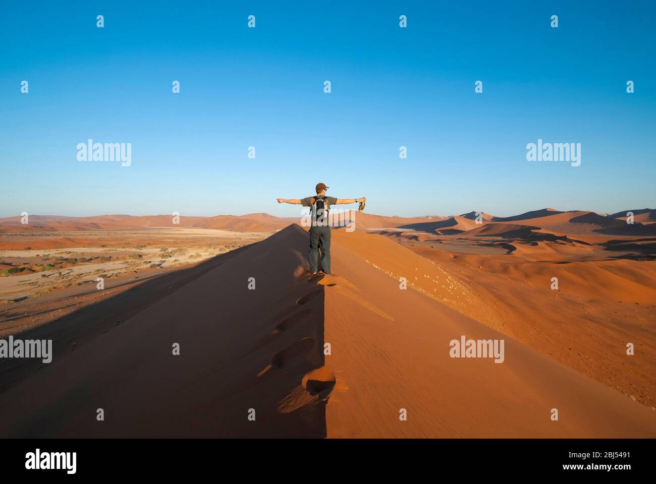 Ein einzelner Mann, der bei Sonnenuntergang in der Namid-Wüste in Sossusvlei, Namibia, Afrika, auf einer Sanddüne mit ausgestreckten Armen unterwegs ist. Querformat. Stockfoto