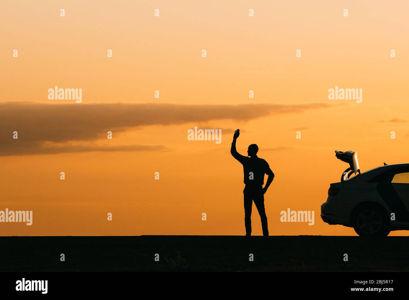 Der männliche Fahrer hob die Hand und wartet auf Hilfe, sein Auto kann sich nach einem Ausfall bei Sonnenuntergang, Seitenansicht, Kopierraum nicht weiter bewegen. Abstimmungen auf der Straße Stockfoto