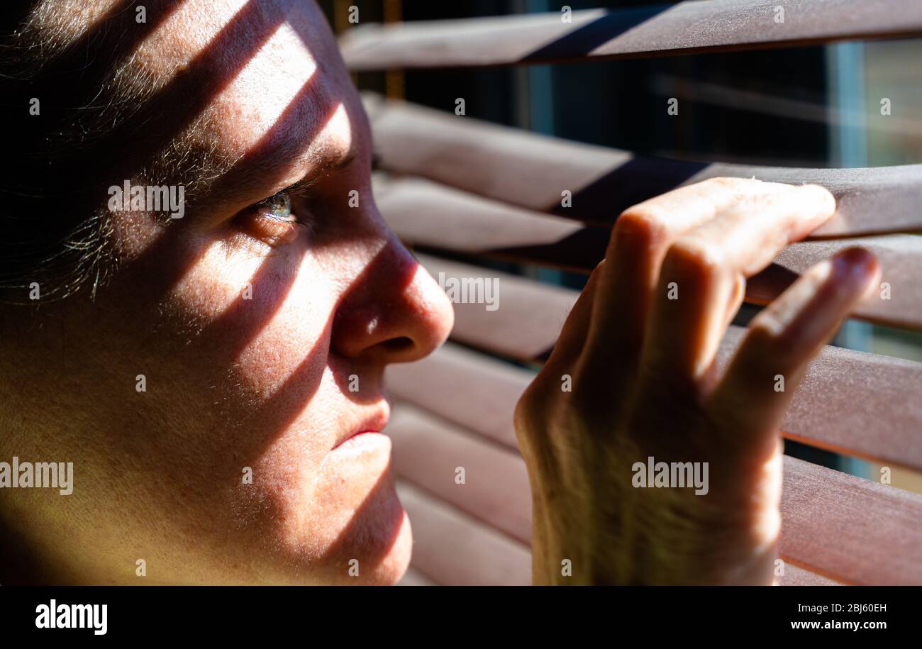 Erwachsene Frau mit blauen Augen, die einen hölzernen Vorhang mit ihren Fingern öffnet, um durch das Fenster zu schauen, während das Sonnenlicht Sonne und Schatten auf ihrem Gesicht erzeugt Stockfoto