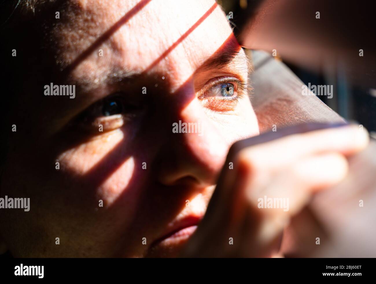 Erwachsene Frau mit blauen Augen, die einen hölzernen Vorhang mit ihren Fingern öffnet, um durch das Fenster zu schauen, während das Sonnenlicht Sonne und Schatten auf ihrem Gesicht erzeugt Stockfoto