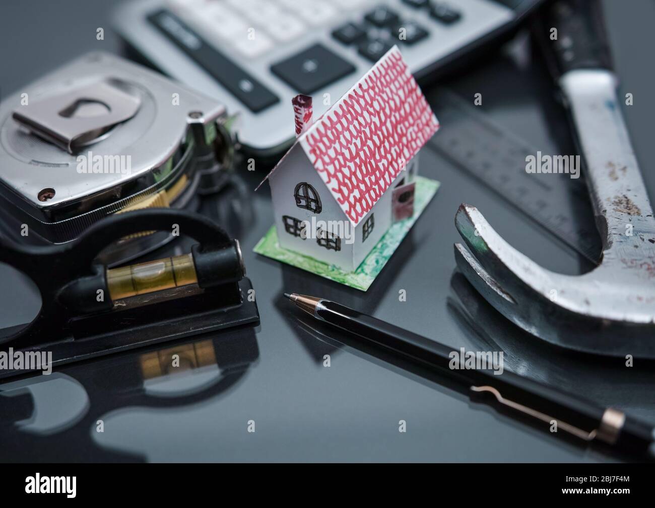Um ein winziges Papierhaus herum haben wir: Einen Hammer, einen Taschenrechner, ein Maßband, eine Vintage-Blase und einen Architektenstift. Stockfoto