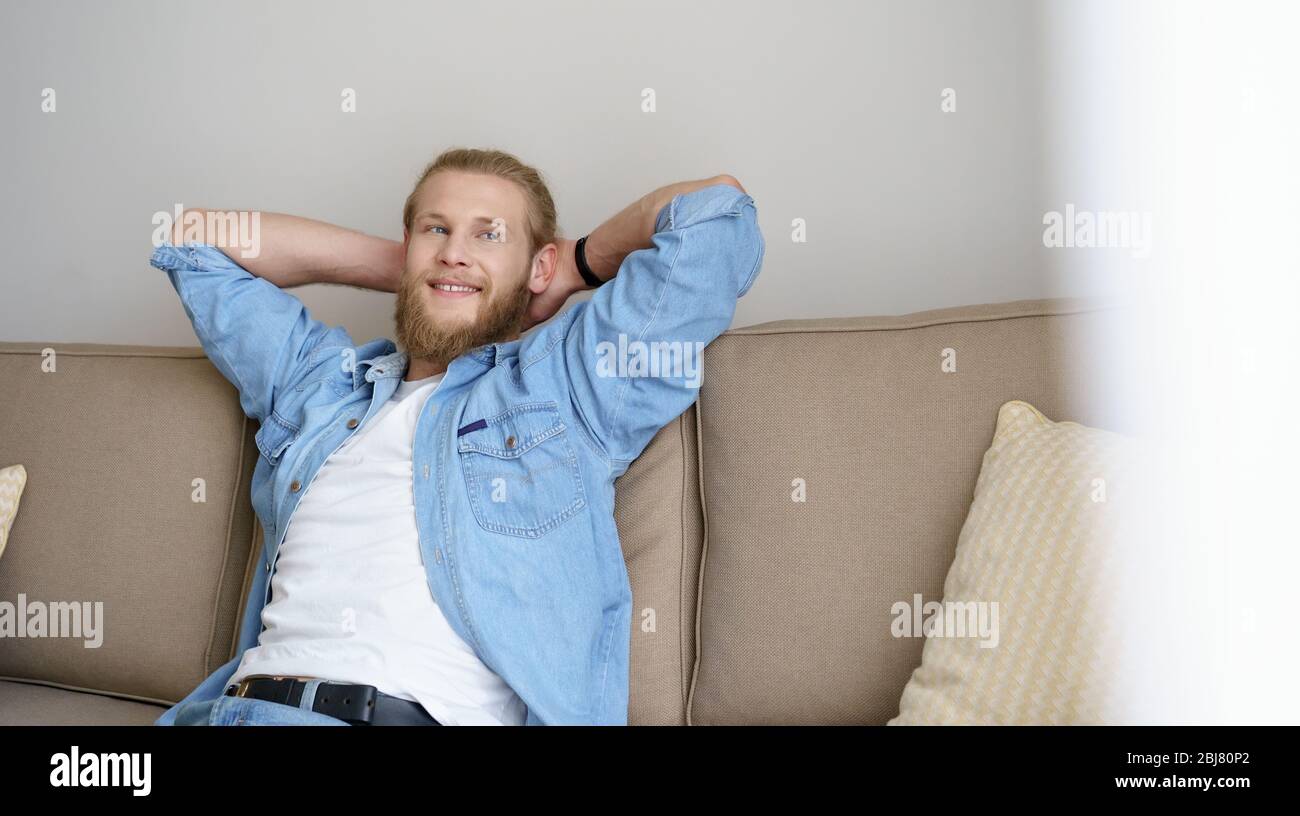 Lächelnd, entspannter junger Mann, der sich auf einem bequemen Sofa entspannen konnte, das wegschaute. Stockfoto