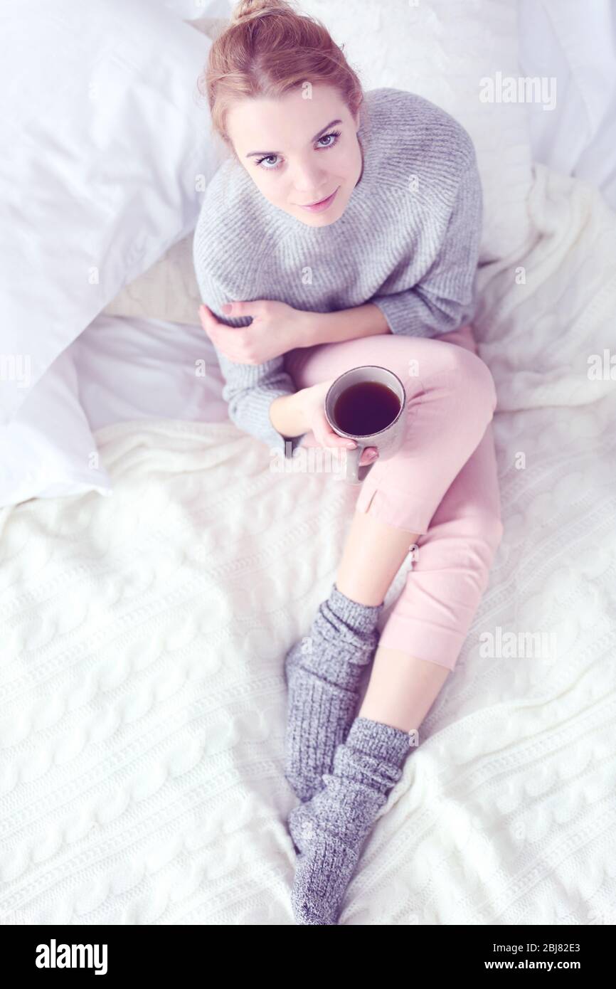 Junge schöne blonde Frau in einem Pyjama und warmen Socken hält eine Tasse Tee, während auf einer weißen Strickdecke sitzen Stockfoto