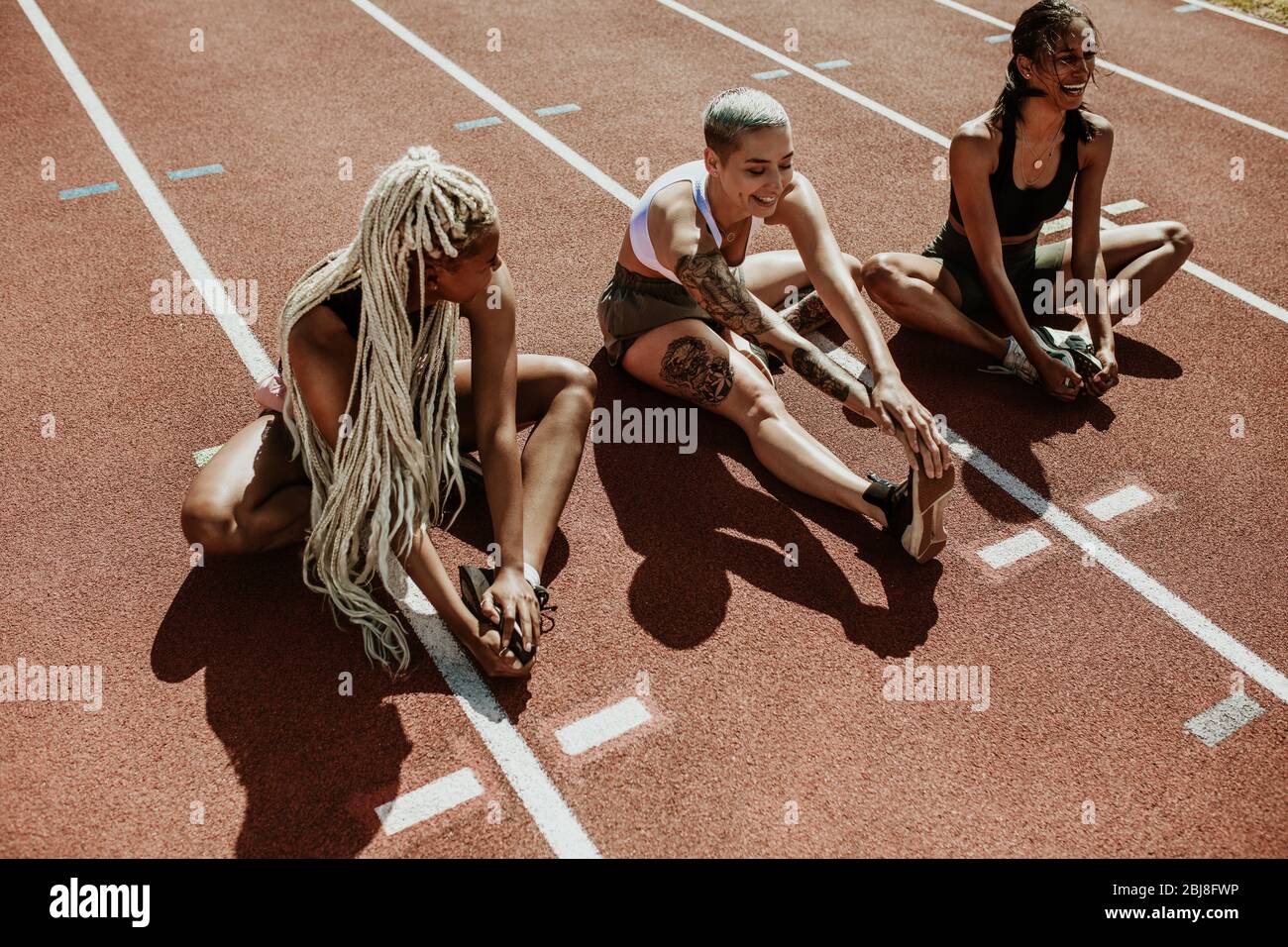 Drei Sportlerinnen sitzen auf einer Laufstrecke und strecken die Beine. Multiethnische Gruppe von Läufern, die im Stadion Aufwärmübungen machen. Stockfoto