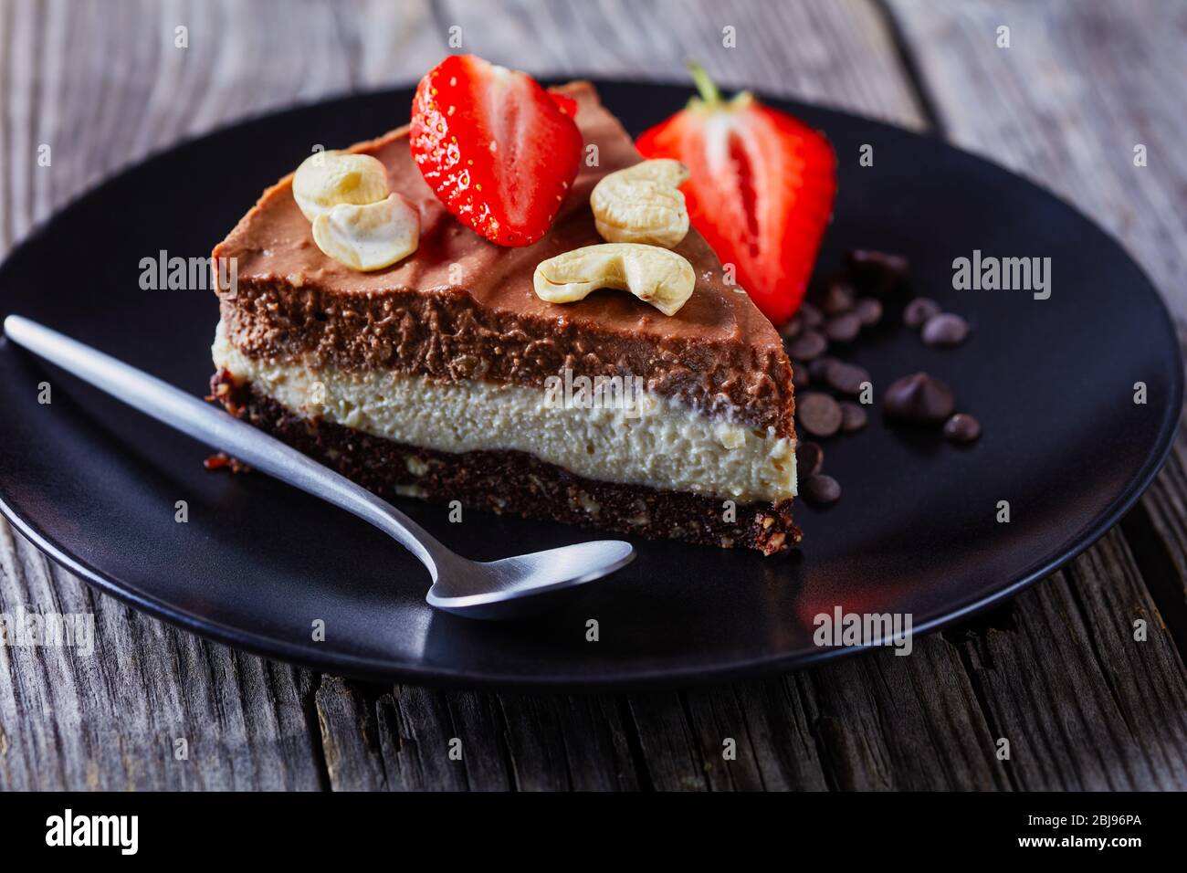 Schokoladen-Tarte aus zwei Schokoladenschichten: Weiß und dunkel mit ungebackenen Kruste aus rohen Datteln, Kakao und Nüssen auf einem schwarzen Teller mit frischen Erdbeeren und c Stockfoto