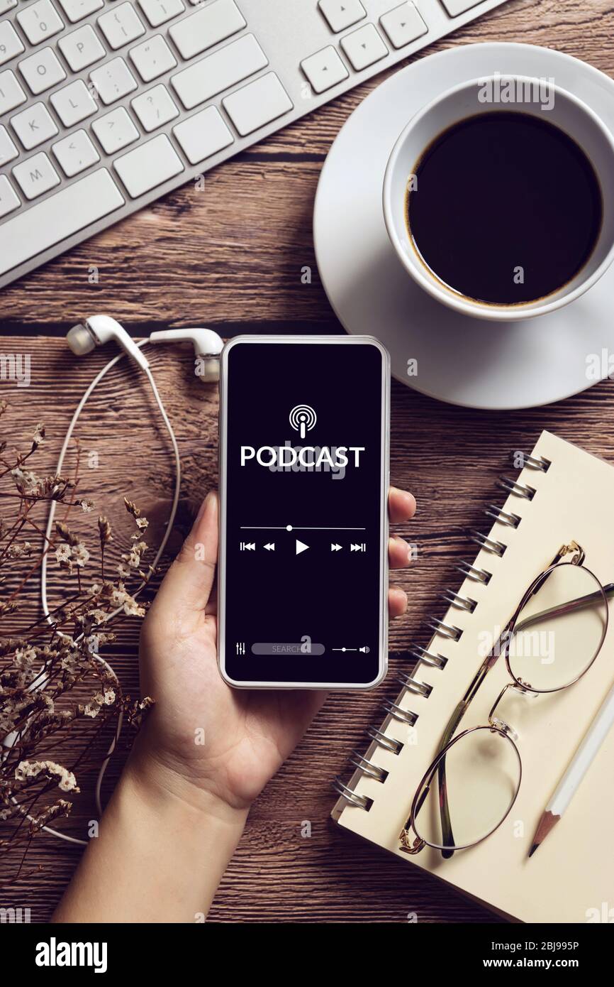 Podcast Audio Content Konzept. Podcast Anwendung auf mobilen Smartphone-Bildschirm auf Holztisch mit Kaffeetasse, Kopfhörer, Brille, Notebook Stockfoto