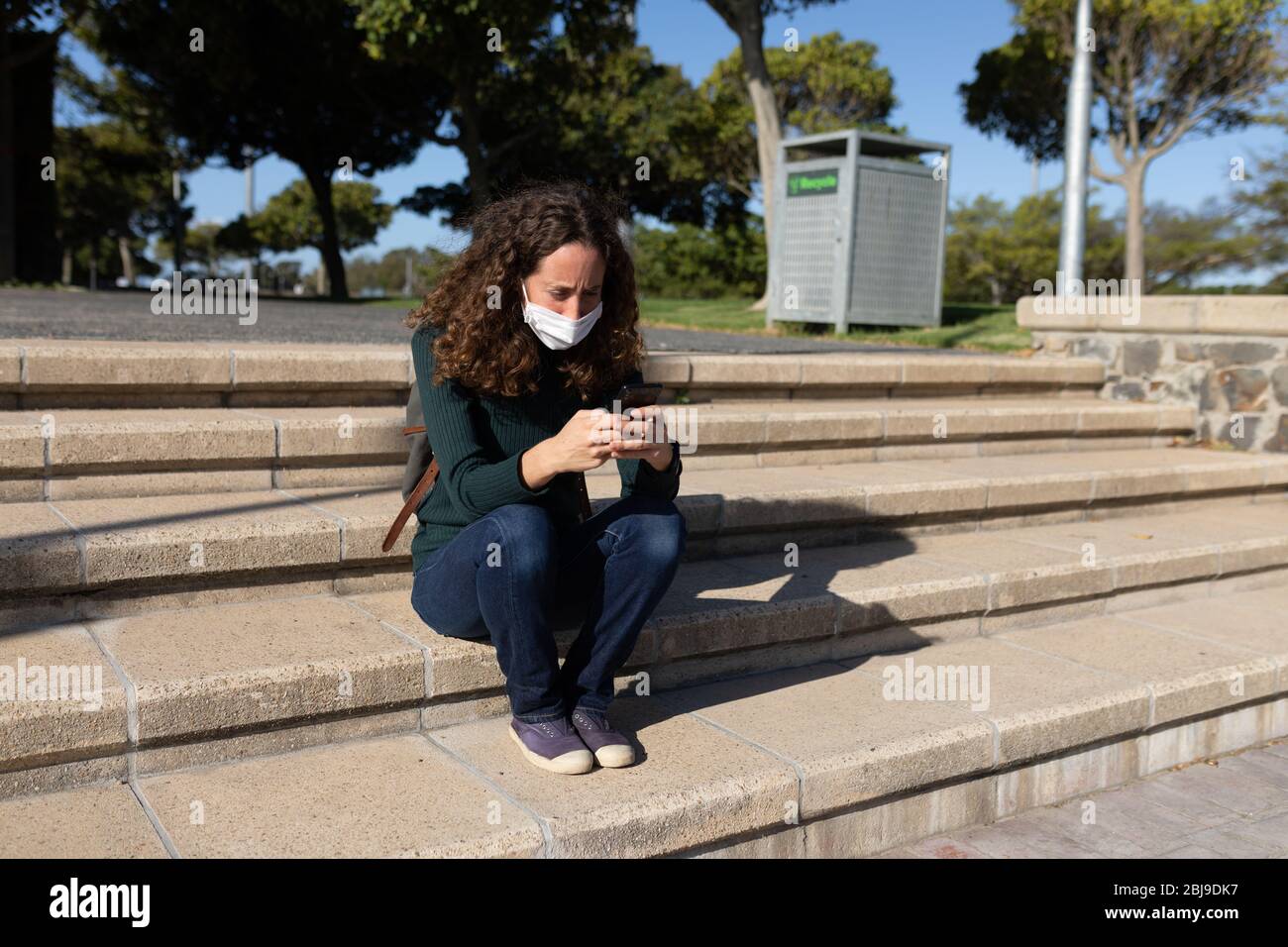 Kaukasische Frau, die eine Schutzmaske auf den Straßen trägt, sitzt und ihr Telefon benutzt Stockfoto