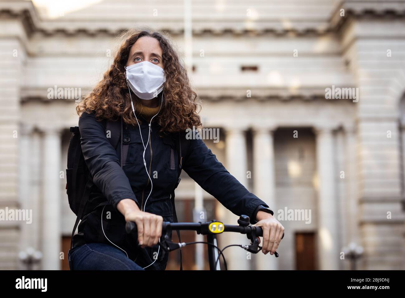 Kaukasische Frau mit einer Schutzmaske auf ihrem Fahrrad, trägt Kopfhörer in den Straßen Stockfoto
