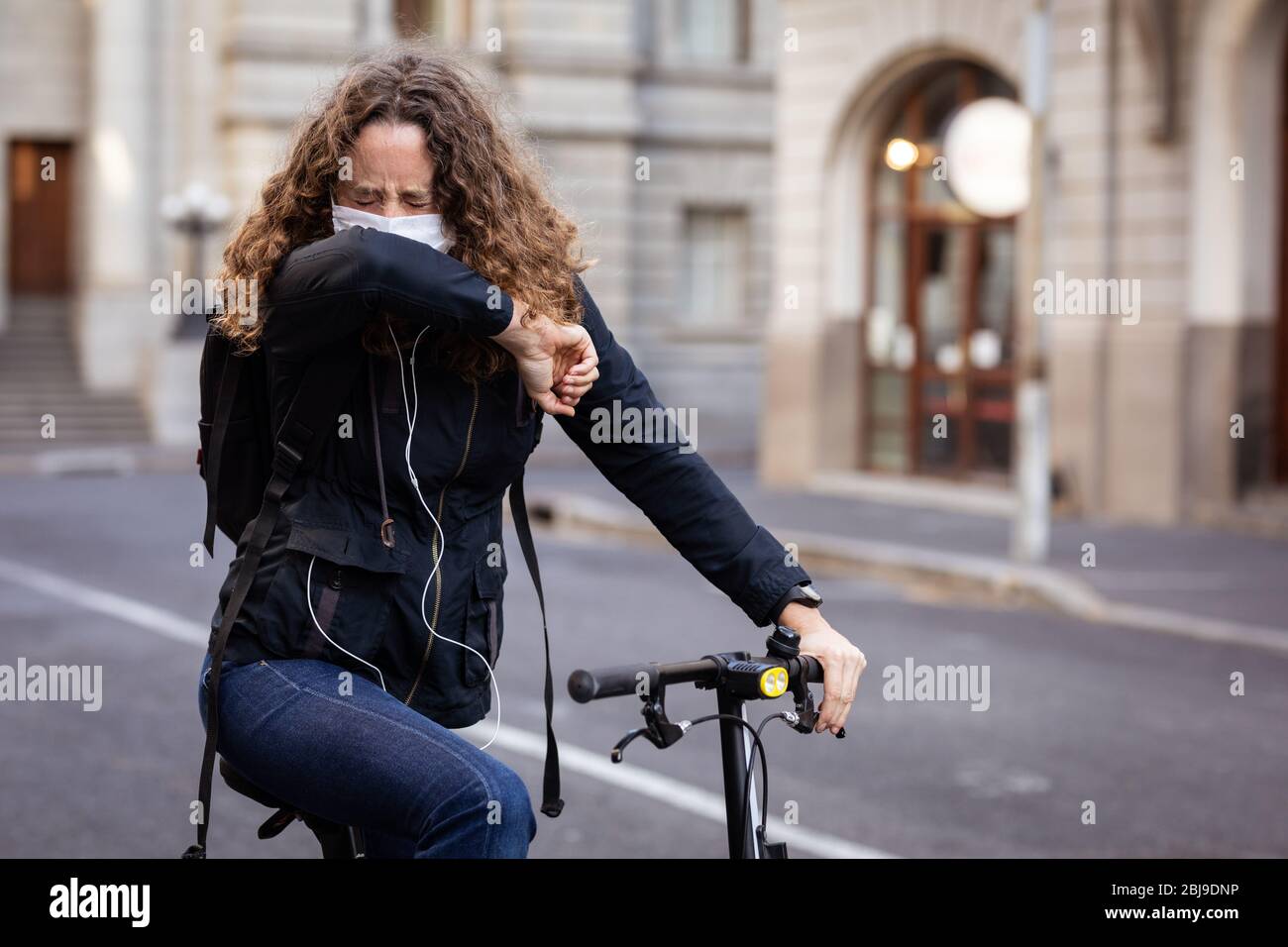 Kaukasische Frau, die eine Schutzmaske auf ihrem Fahrrad trägt, hustet und trägt Kopfhörer in den Straßen Stockfoto